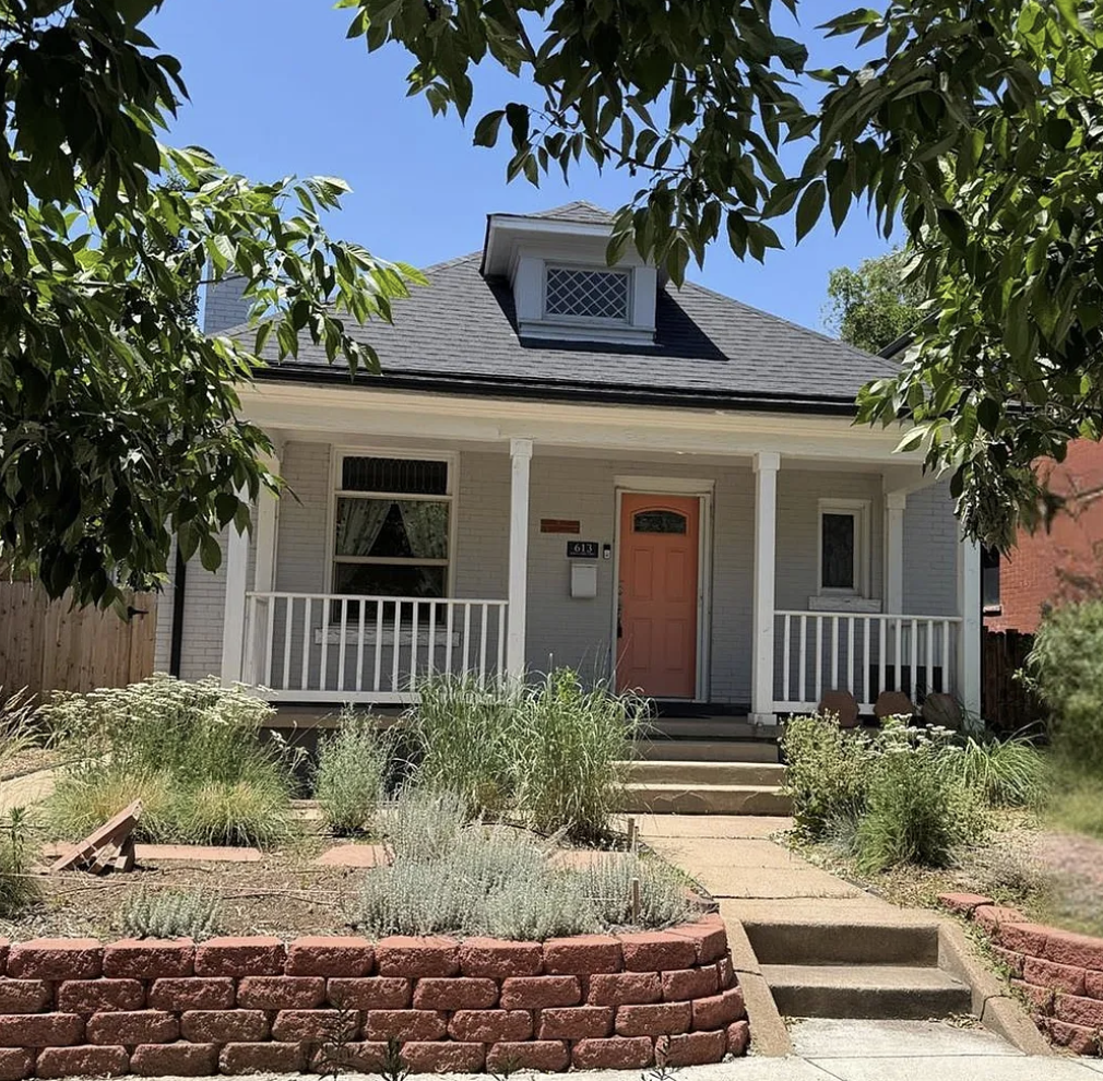 Front view of a house with a white exterior, orange front door, and a small porch. There are three steps leading up to the porch, with a garden of plants and bushes in front. The house has a dormer window on the roof, and there are trees framing the scene against a clear blue sky.
