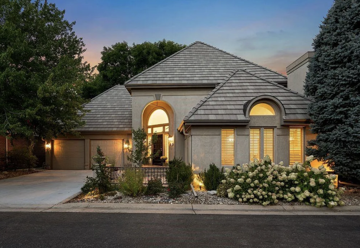 A modern two-story house with a tiled roof, large illuminated windows with shutters, and a landscaped front yard with bushes and trees at dusk.