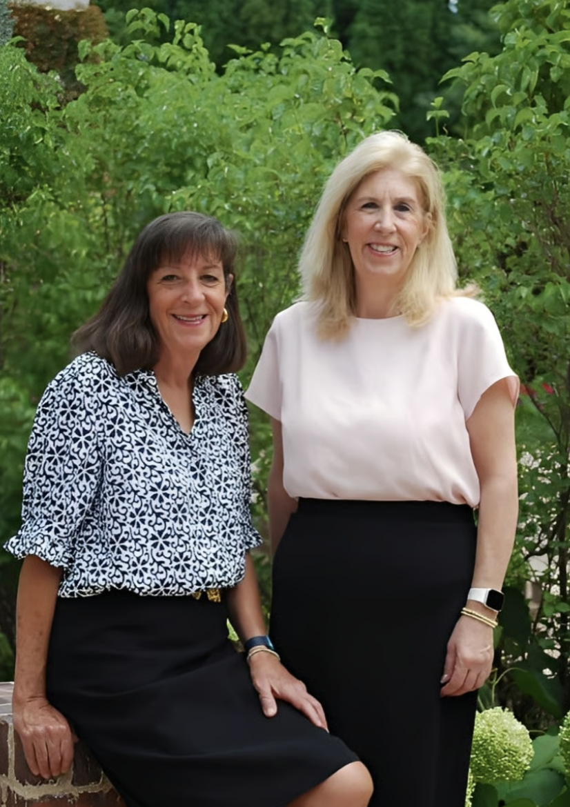 Two women standing outdoors in front of green foliage, smiling at the camera.