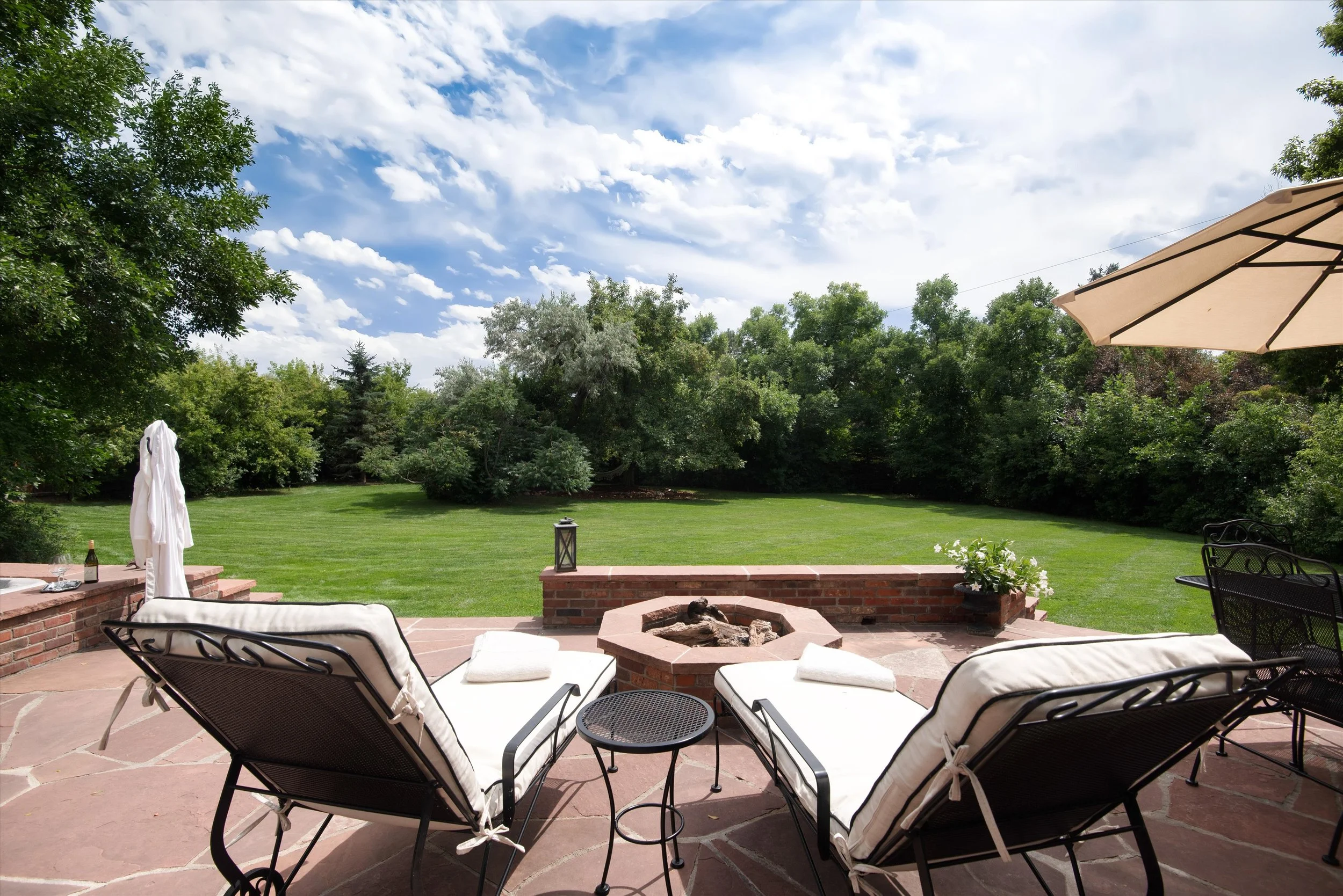 A backyard patio with black metal lounge chairs with white cushions, a small round black table, a brick fire pit, and a brick ledge with a lantern and white towels. Beyond the patio is a lush green lawn and a line of trees under a partly cloudy blue sky.