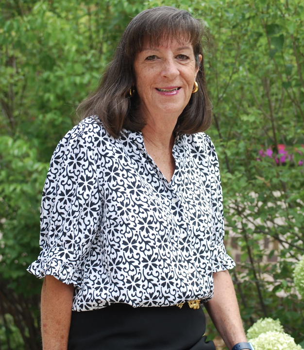 A middle-aged woman with shoulder-length brown hair smiling outdoors, wearing a patterned black and white blouse and gold earrings, with green foliage in the background.
