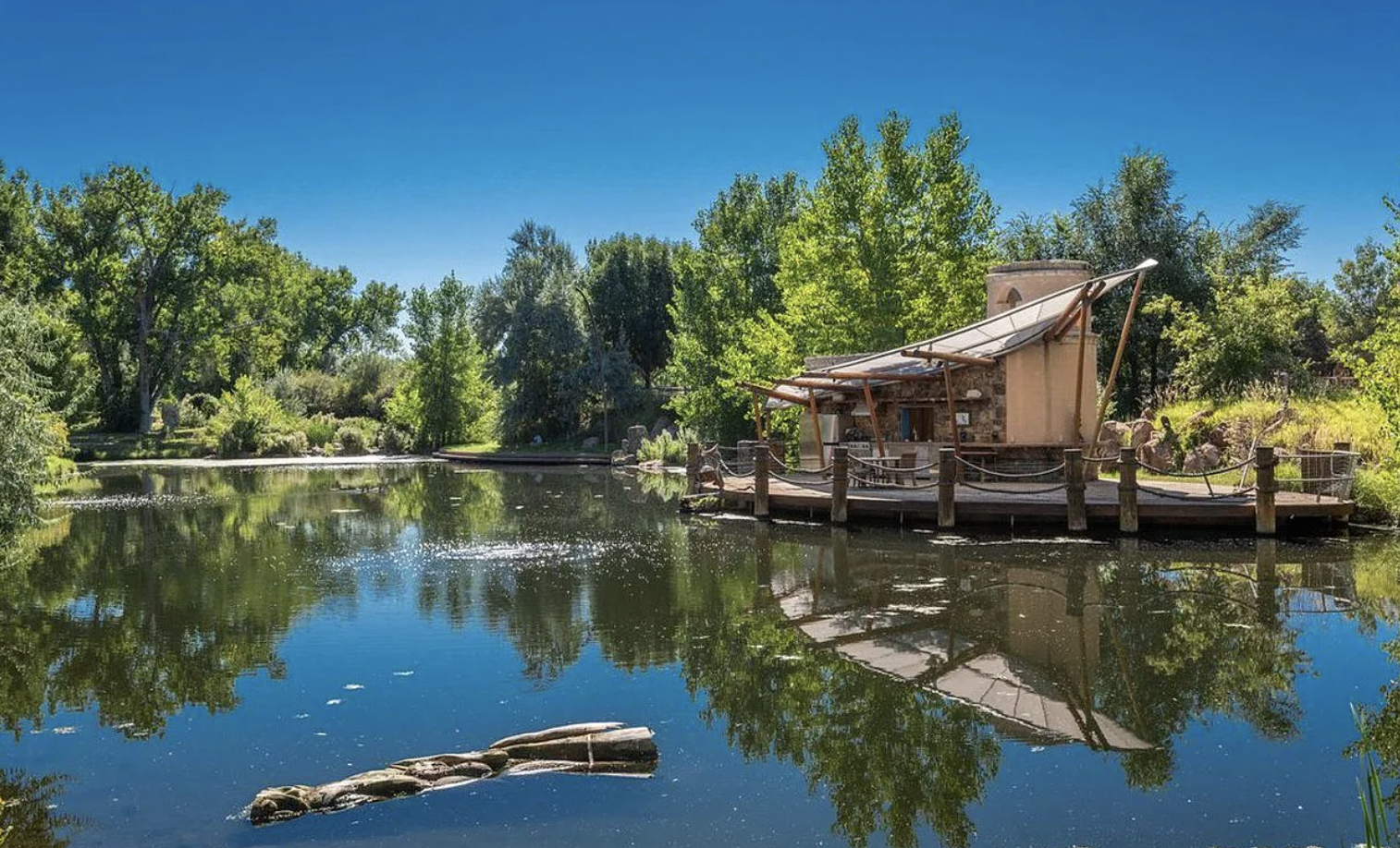 A peaceful pond surrounded by lush green trees with a modern, open-air pavilion featuring a canopy over a deck on the right side, reflecting in the water under a clear blue sky.