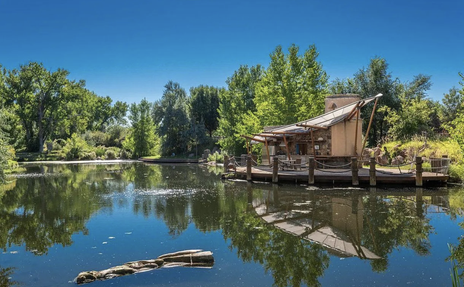 A peaceful scene of a lake surrounded by green trees under a clear blue sky, with a small house or hut built on the edge of the water, featuring a slanted roof and a wooden deck with a rope railing. The water reflects the trees and the house.