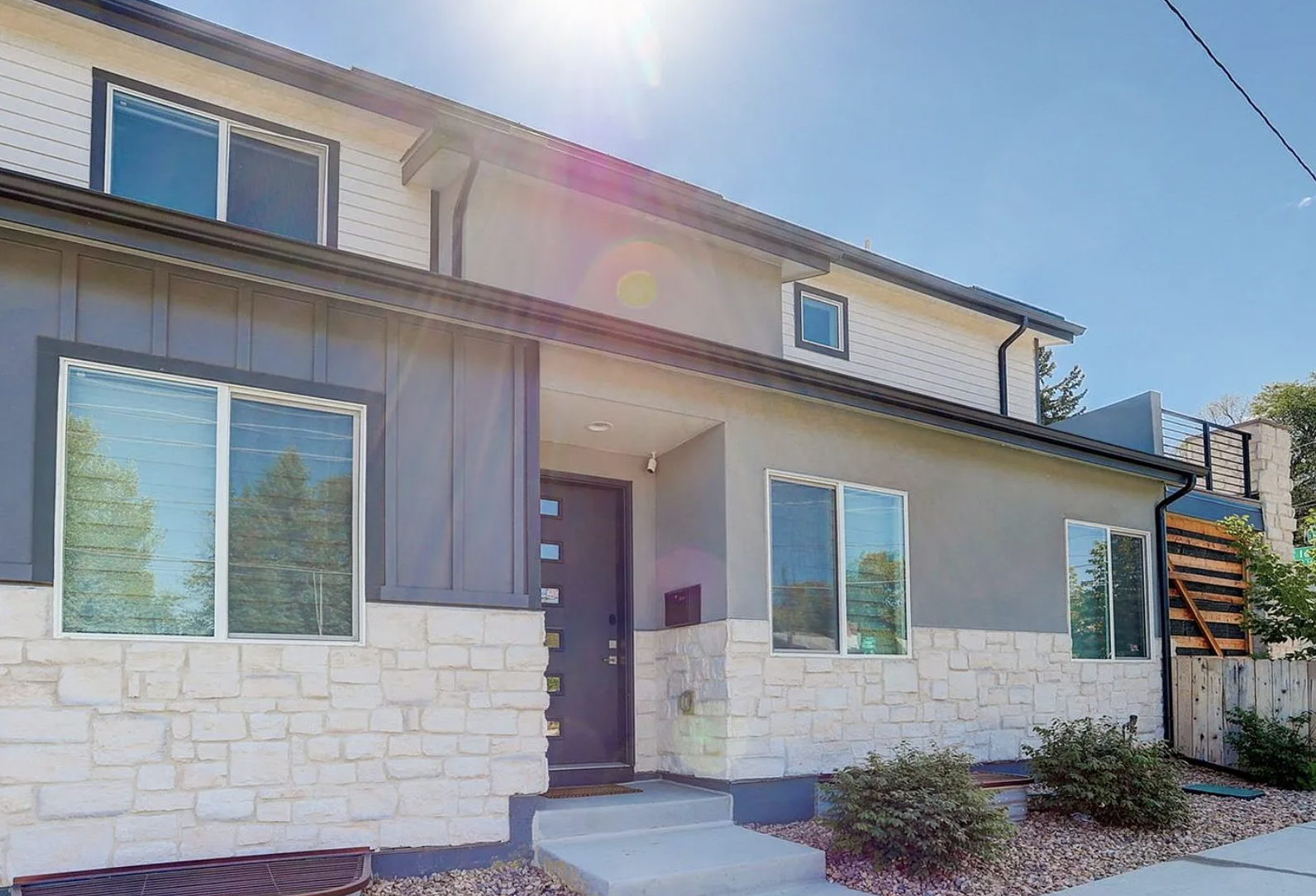 Modern two-story house with stone and siding exterior, large windows, and a front door with glass panels, under a clear blue sky with sunlight.