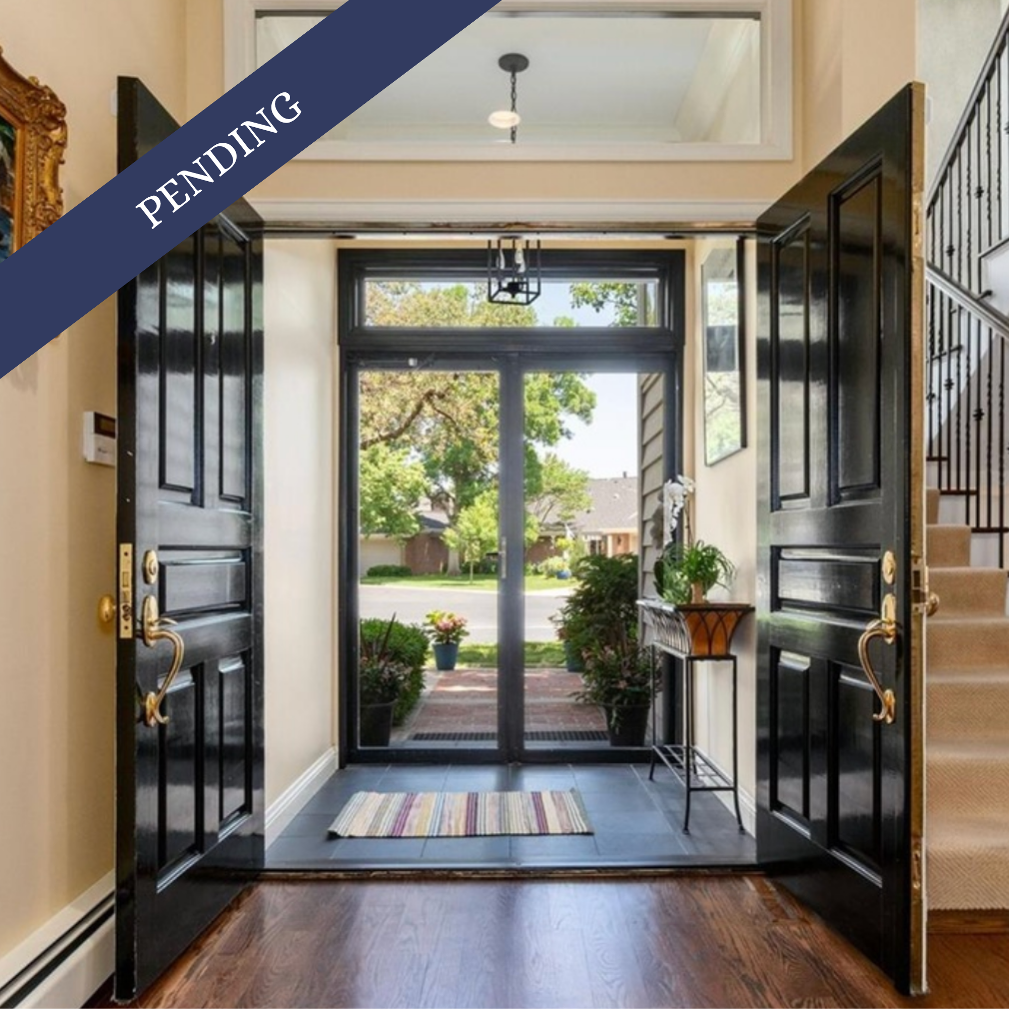 Open front door leading to a welcoming entryway with a view of a sunny street, a striped rug on a wooden floor, potted plants, and a chandelier hanging from the ceiling.