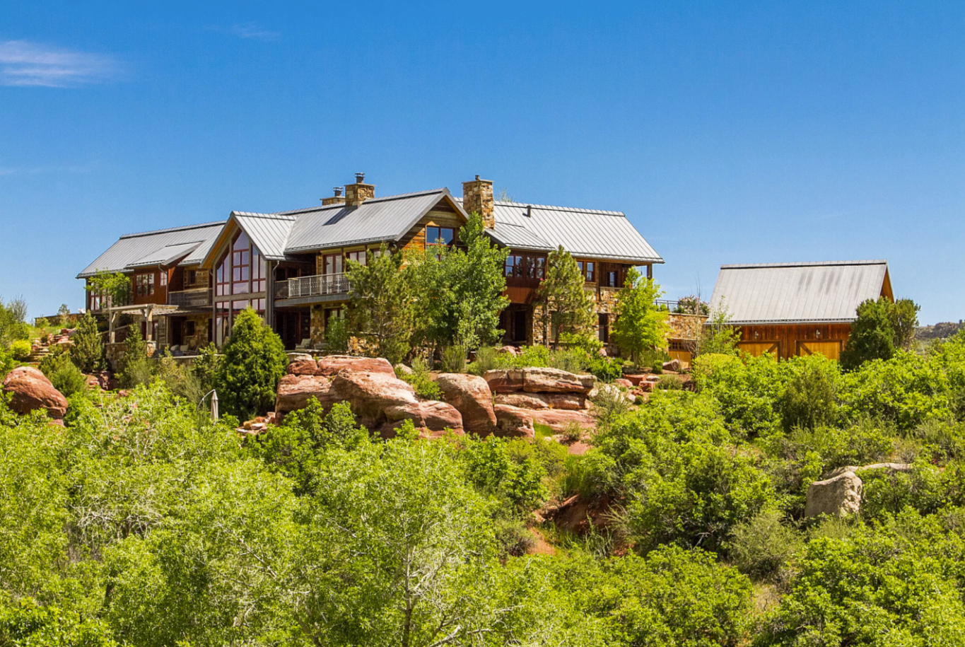 A large house built on a hillside surrounded by green trees and rocks, under a bright blue sky.