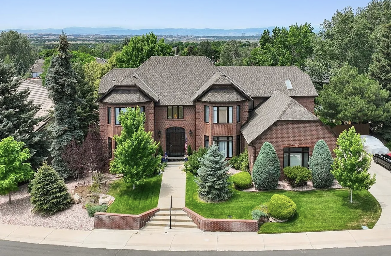 Large brick house with a well-manicured green lawn, various trees, and shrubs in the front yard. The house has multiple gabled roofs, large windows, and a central entrance with a pathway leading to the sidewalk.