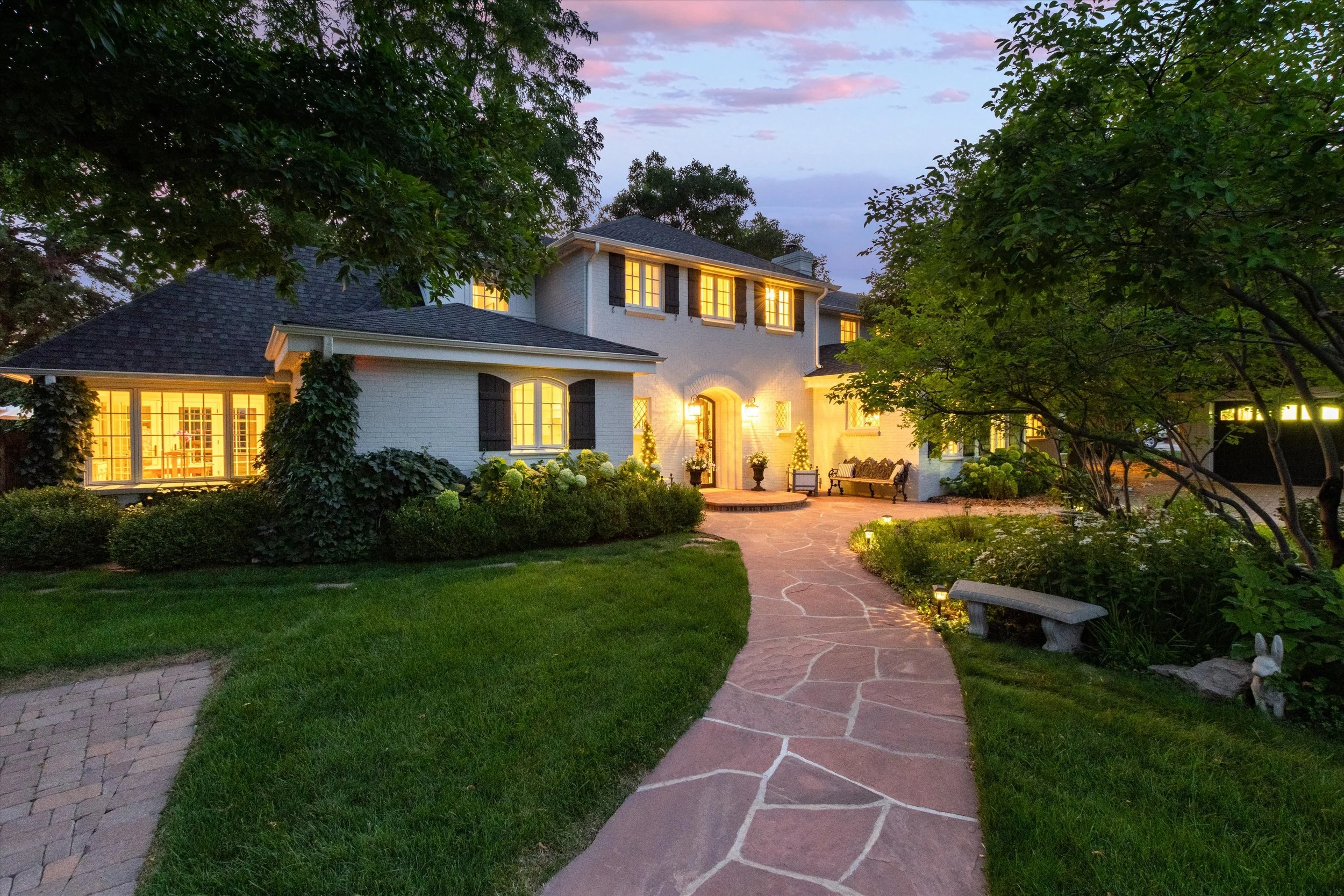 A large two-story house illuminated from within, with trees and greenery surrounding a stone pathway leading to the front door, under a twilight sky.