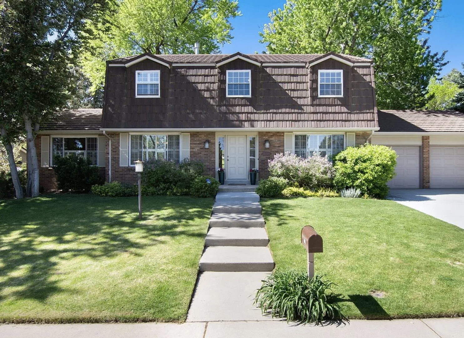 A two-story house with a brick lower level and dark brown shingle upper level, surrounded by a green lawn, trees, and shrubs. There is a concrete walkway leading to the front door, a mailbox in the yard, and two garage doors on the right.