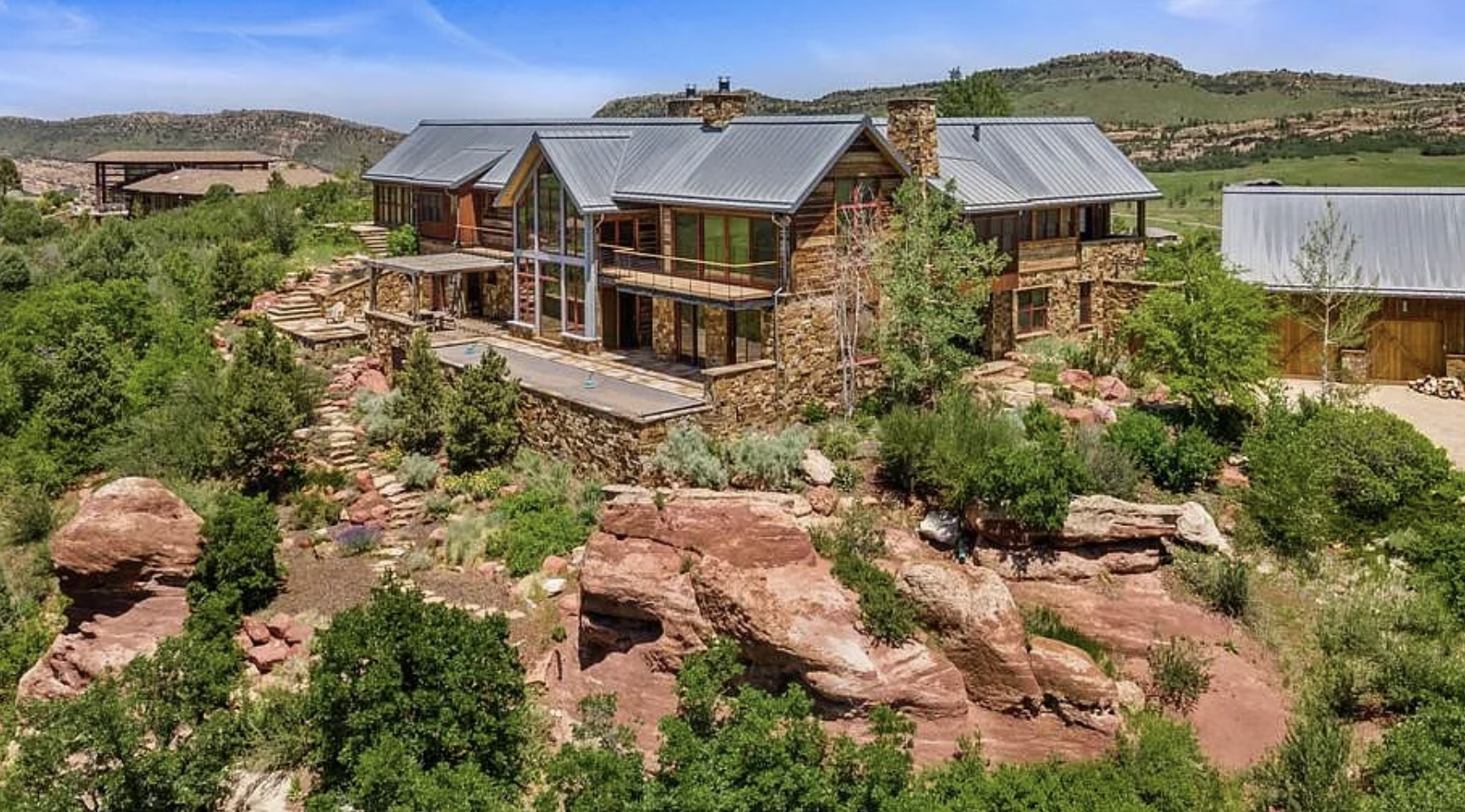 A modern house built with wood and stone, situated on rocky terrain with a landscaped garden and lush greenery, surrounded by hills under a clear blue sky.