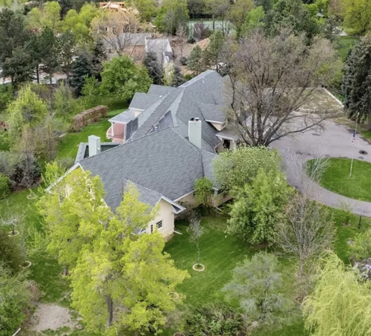 Aerial view of a suburban house with a large gray roof surrounded by trees and a manicured lawn, with a curved driveway and a small parking area.