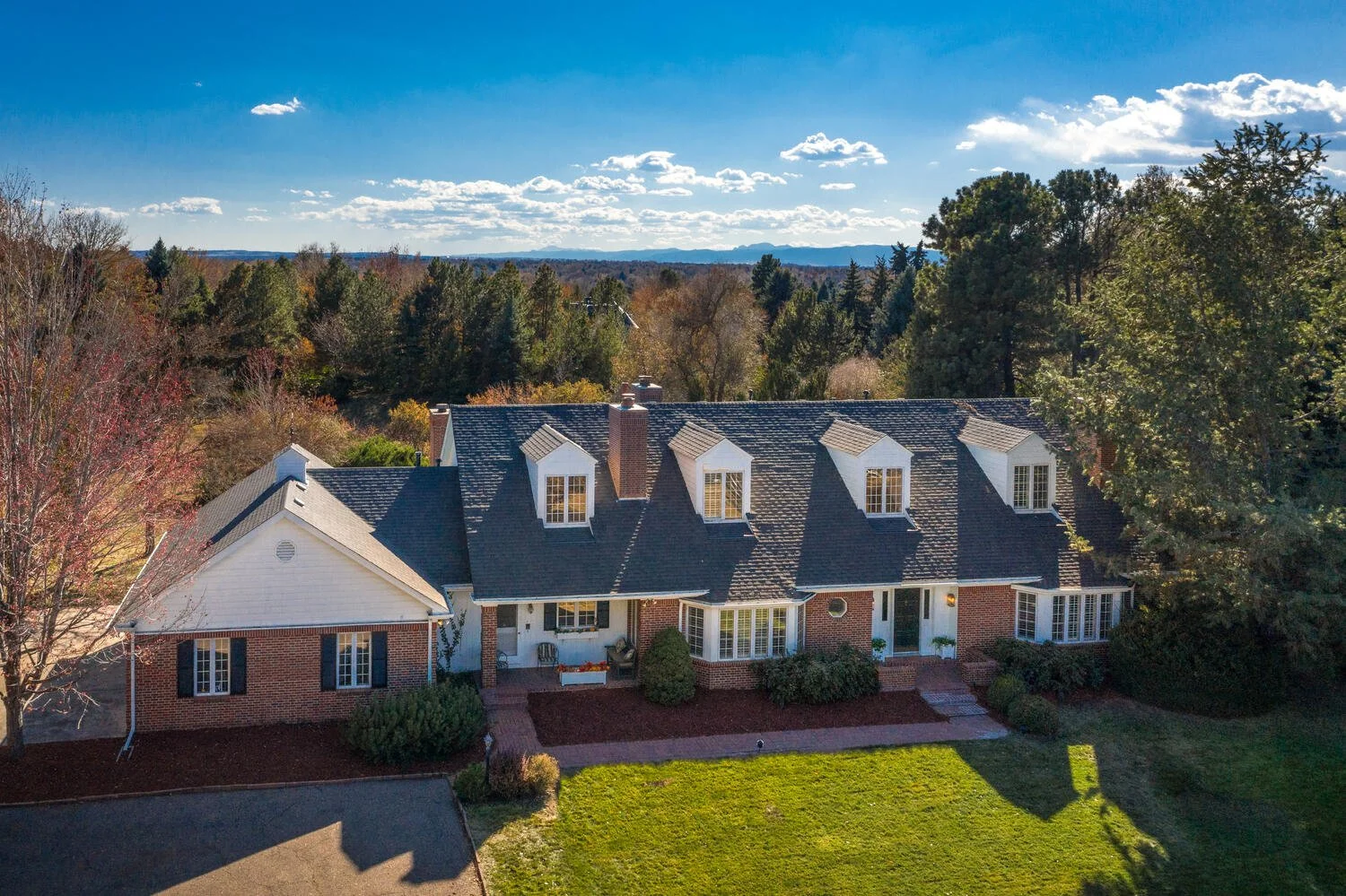A large two-story house with a brick and white exterior, dark shingle roof, and five dormer windows, surrounded by a green lawn and trees with a background of woods and mountains under a blue sky with some clouds.