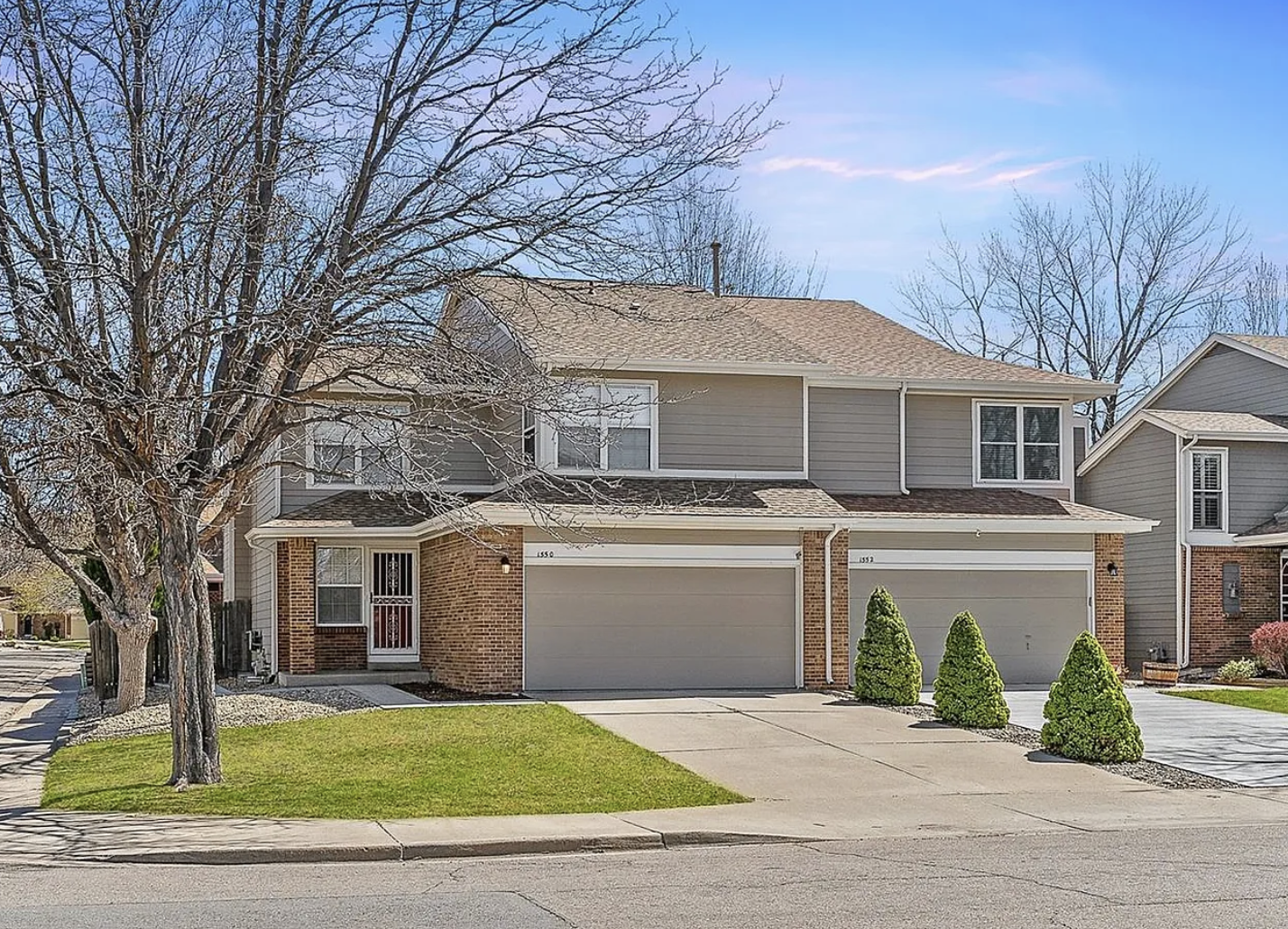 A two-story suburban house with gray siding, brick accents, two attached garages, a front lawn with small bushes, a large leafless tree, and neighboring houses under a clear blue sky.