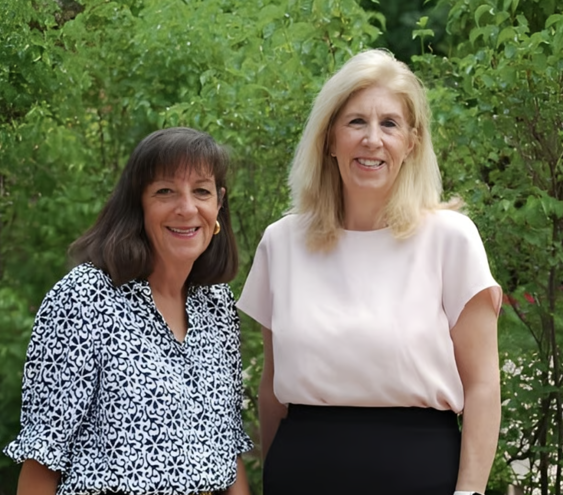 Two women standing outdoors in front of green bushes, smiling at the camera.