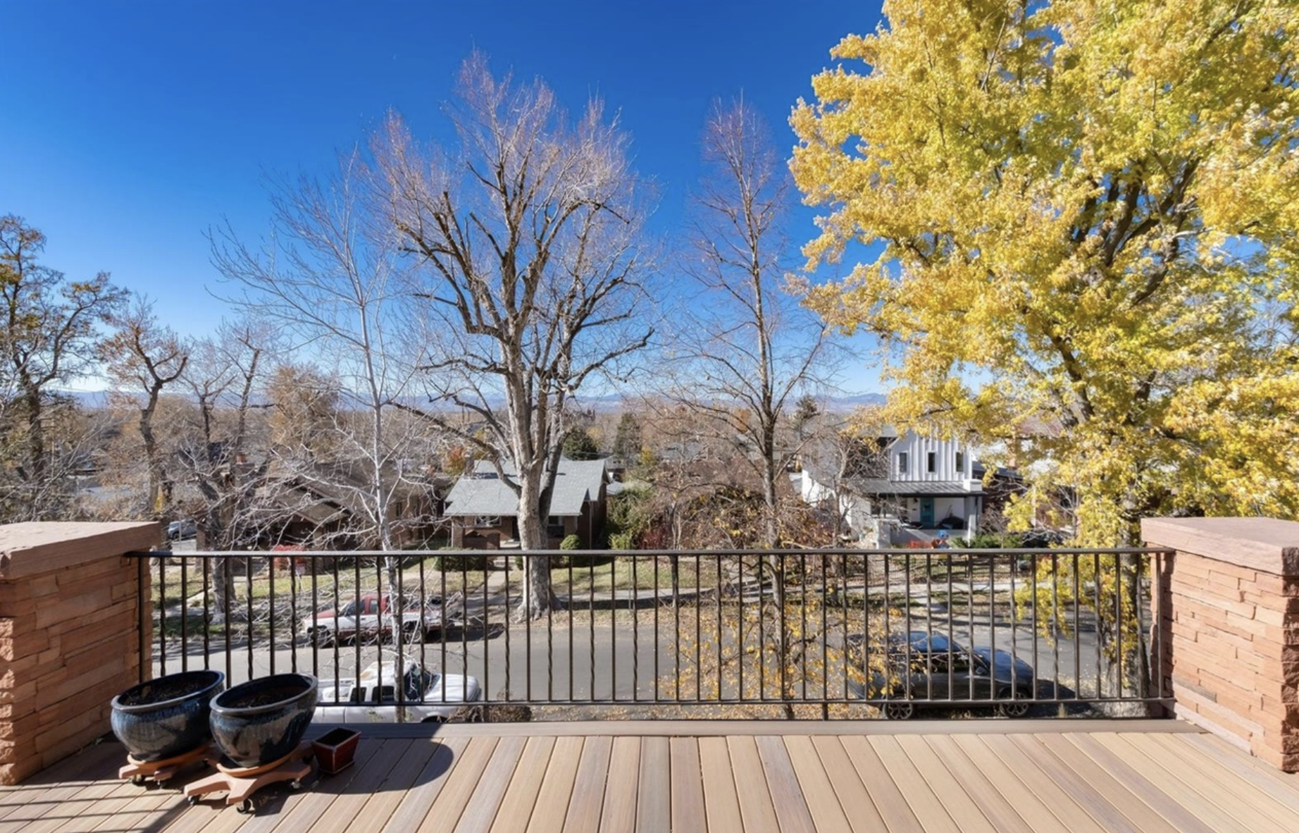 View from a balcony with wooden flooring and a black metal railing. In the foreground, there are three empty black plant pots. Beyond the balcony, there are parking cars, leafless trees, and trees with yellow foliage. Residential houses with rooftops and balconies are visible, along with a clear blue sky.