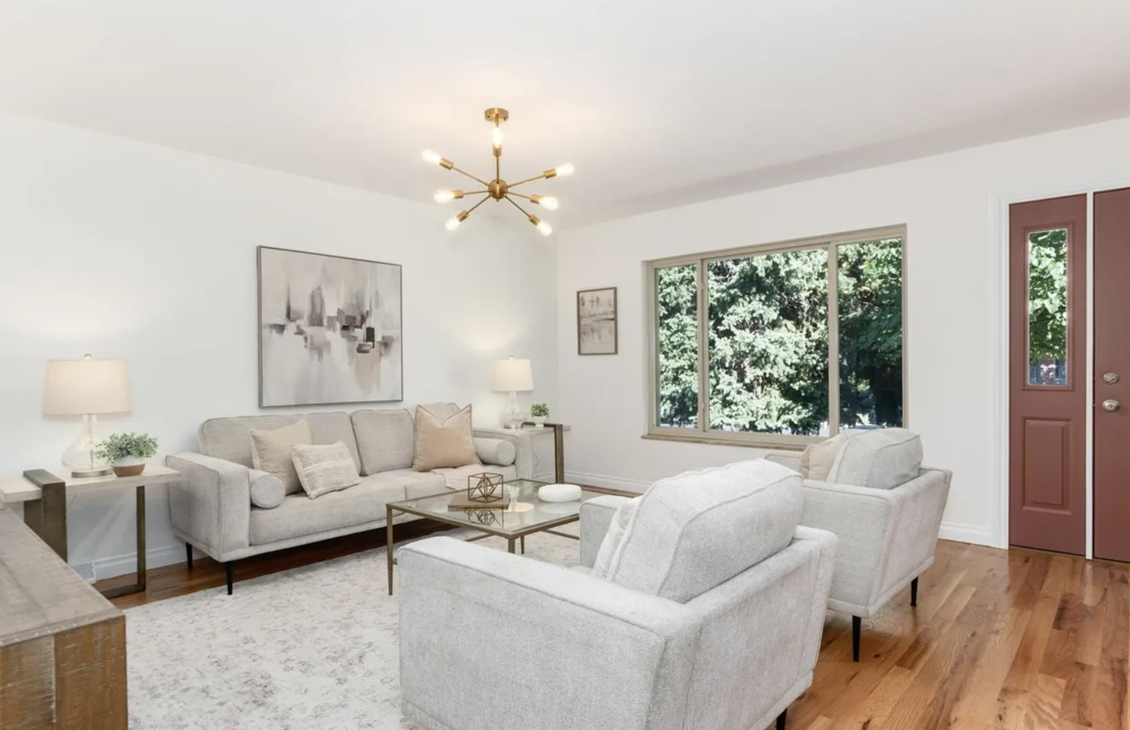 Living room with white walls, beige furniture, wooden flooring, and large windows showing trees outside.