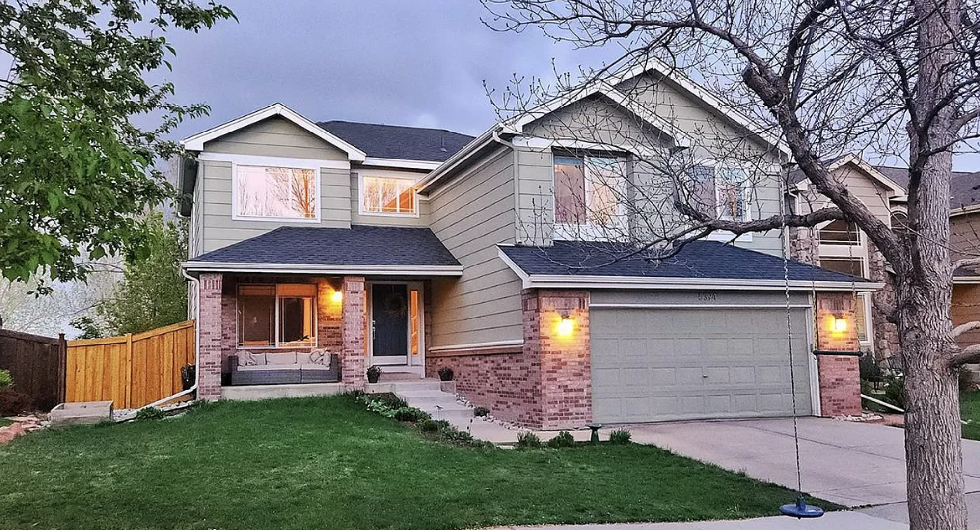 A two-story suburban house with a gray roof, green siding, brick accents, a small front porch with a cushioned seating area, and a garage with lighting on. There are trees, a grassy lawn, and a wooden fence on one side. A swing hangs from one tree, and the sky is cloudy.