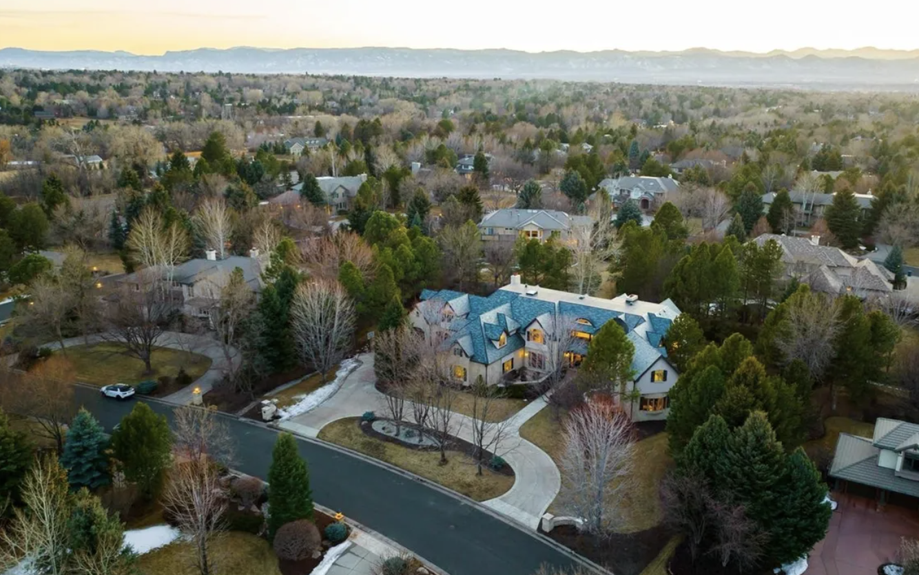 An aerial view of a residential neighborhood with a large house in the foreground, surrounded by trees and other houses, with patches of snow on the ground.