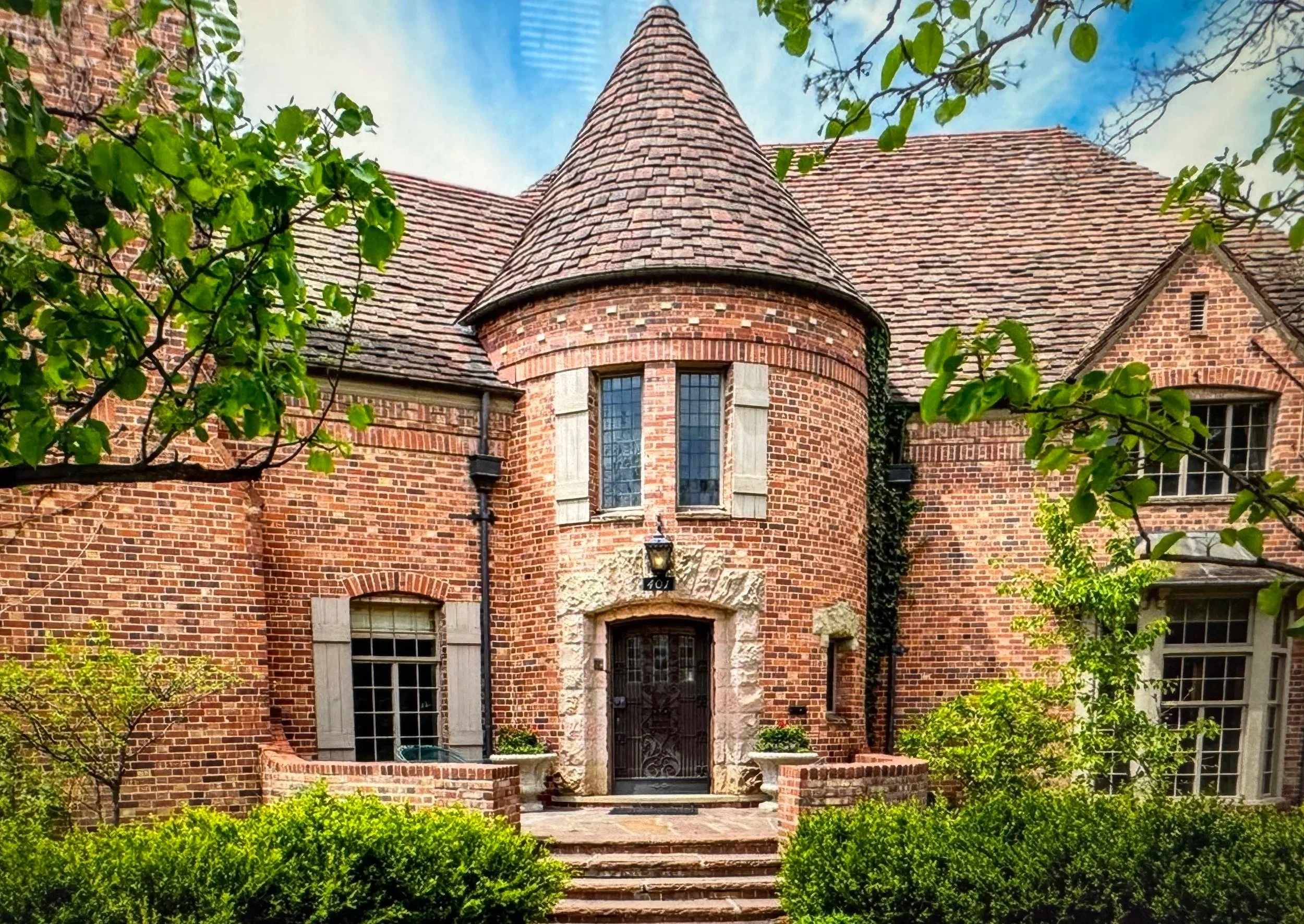 A brick house with a turret and arched windows, surrounded by greenery and trees, with steps leading to the front door.