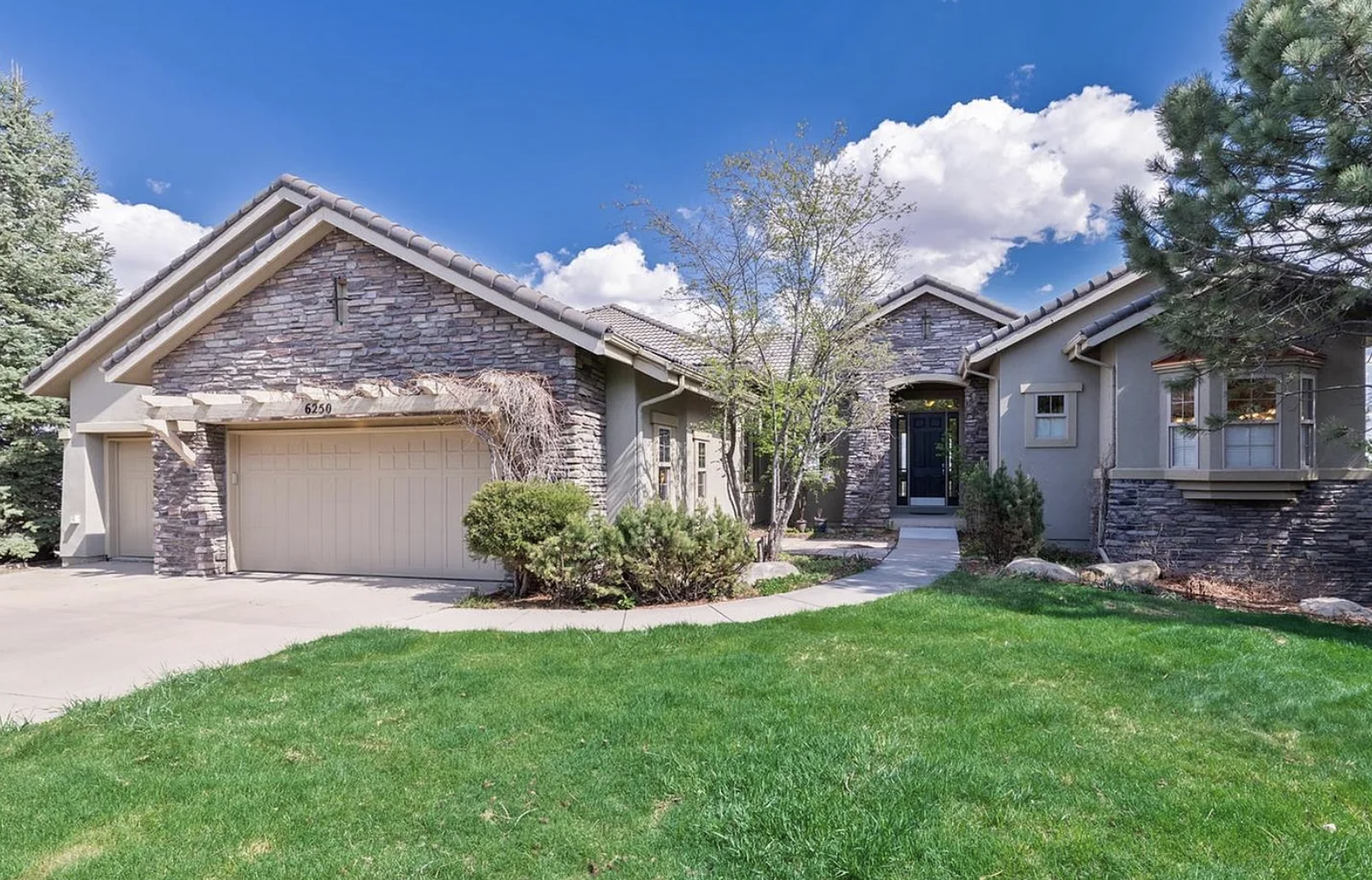 Front view of a modern house with a green lawn, stone accents, garage, and a walkway leading to the front door under a partly cloudy sky.