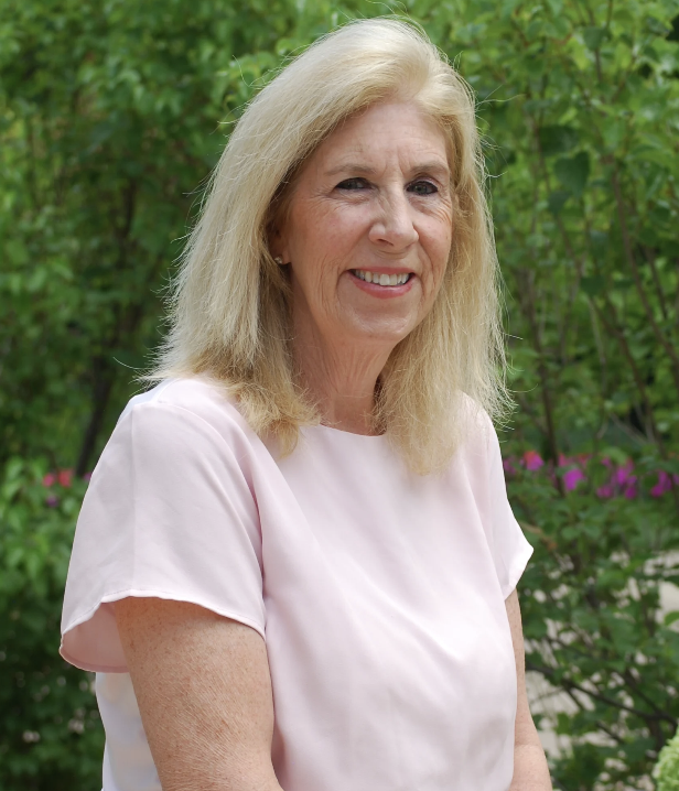 A smiling older woman with blonde hair wearing a light pink blouse standing outdoors with green foliage and pink flowers in the background.