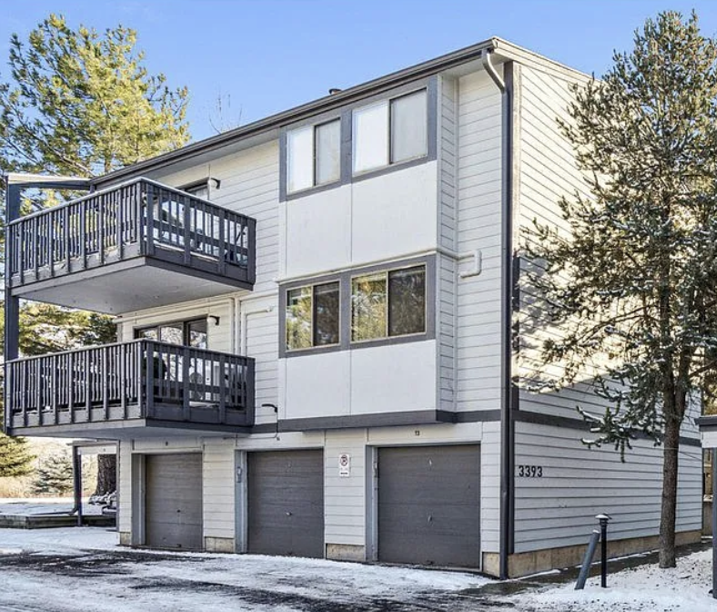 Three-story residential building with a garage on the ground level, balconies on the second and third floors, white exterior, and surrounded by pine trees in winter with snow on the ground.