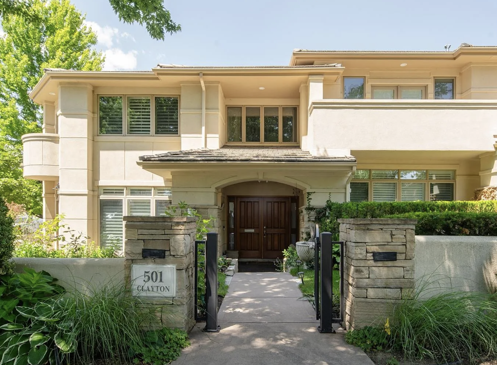 Front view of a modern residential house with a stone and stucco exterior, green shrubs, and a wooden front door, surrounded by a stone mailbox and a small garden.