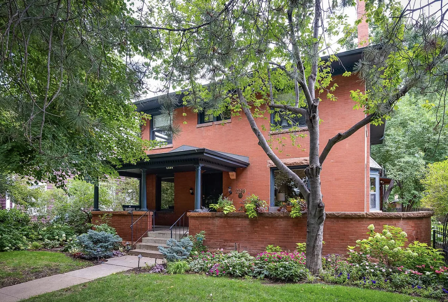 A two-story brick house with a front porch, black roof, and windows, surrounded by a lush garden with trees, shrubs, and colorful flowers.