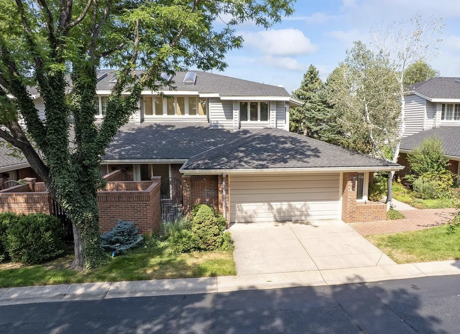 A two-story house with a gray roof, brick and siding exterior, a driveway with a garage, and surrounding trees and greenery. There is a brick wall and garden in front of the house.