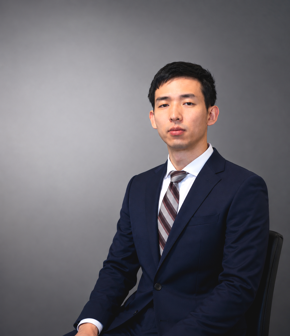 A young man in a navy suit, white shirt, and striped tie sitting on a black chair against a plain gray background.