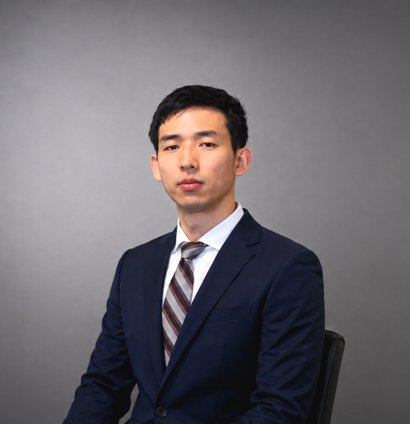 A young Asian man in a navy suit, white shirt, and striped tie, sitting against a gray background, looking at the camera with a neutral expression.