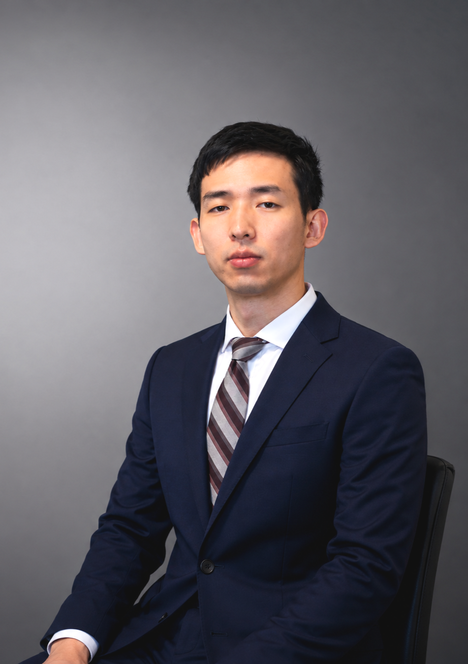 A young man wearing a navy suit, white shirt, and striped tie, seated against a plain gray background.