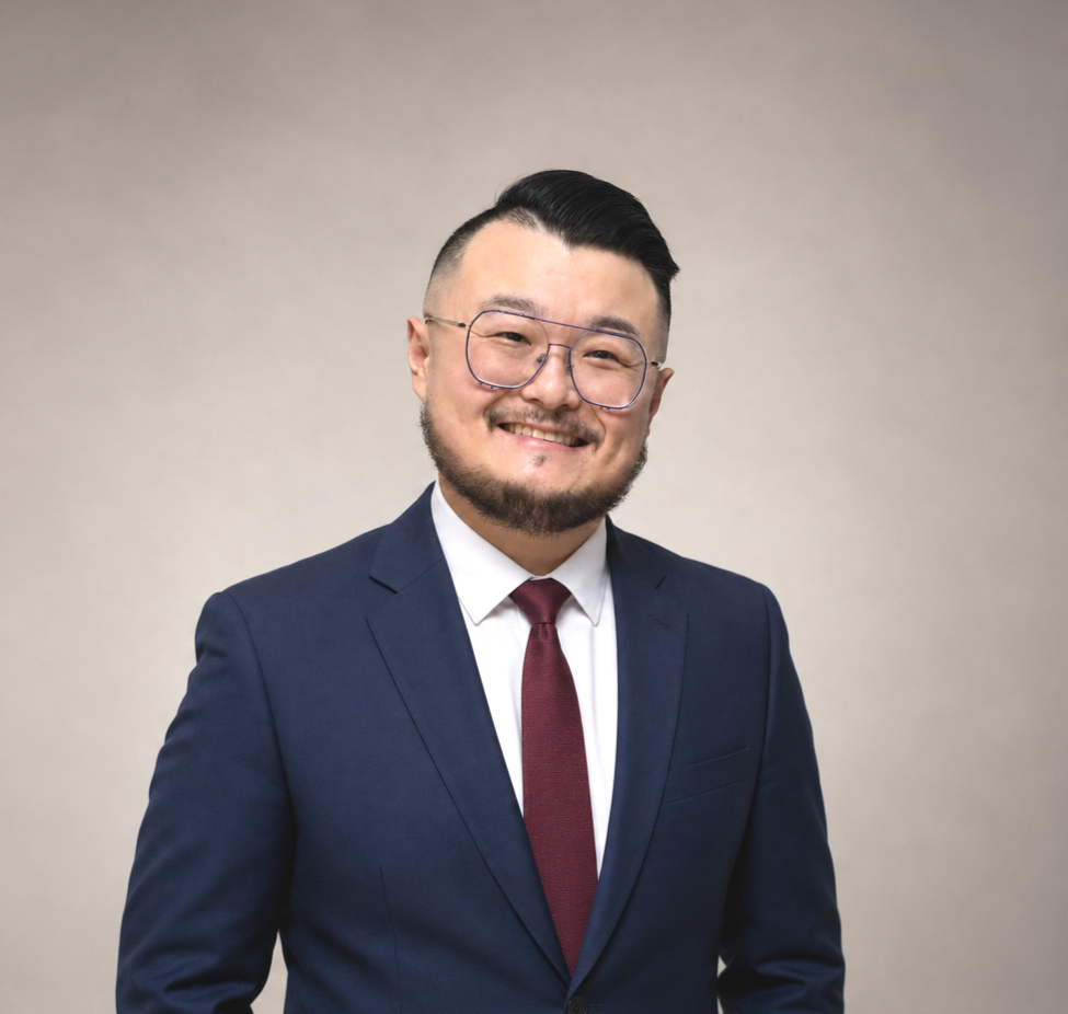 Smiling man with glasses in a suit and tie standing against a plain background.