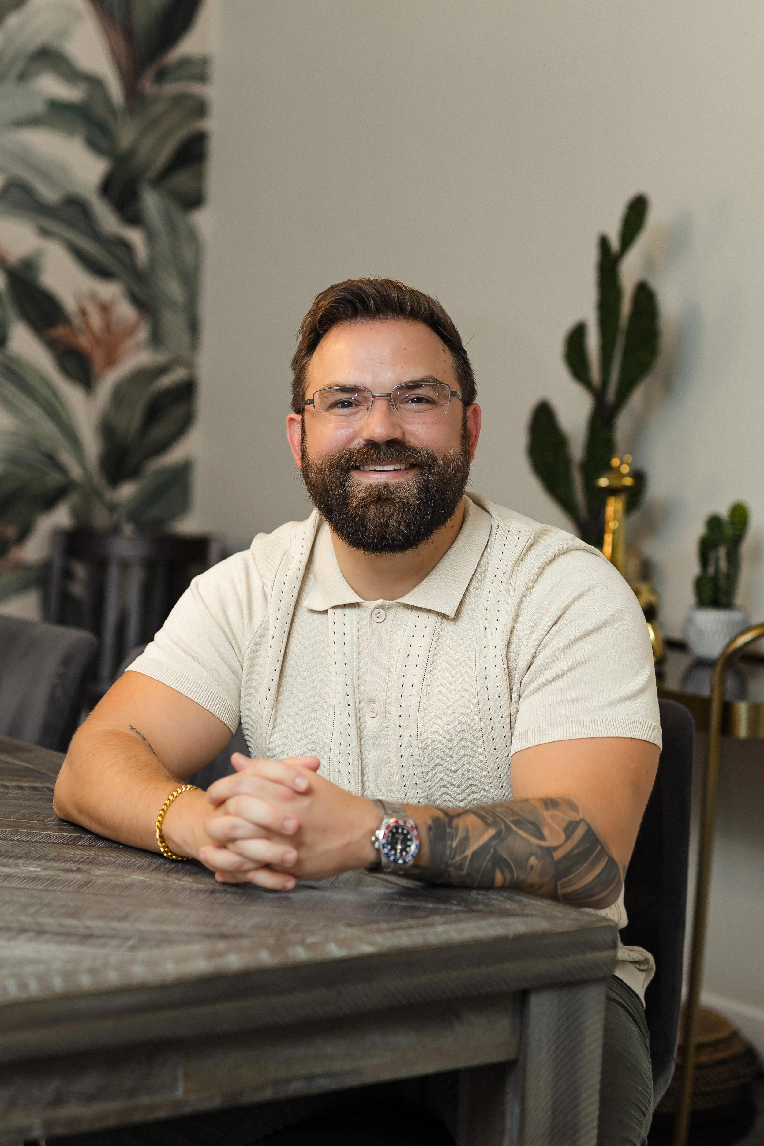 A man with glasses, a beard, and a tattoo on his left arm sitting at a wooden table, smiling at the camera, with plants in the background.