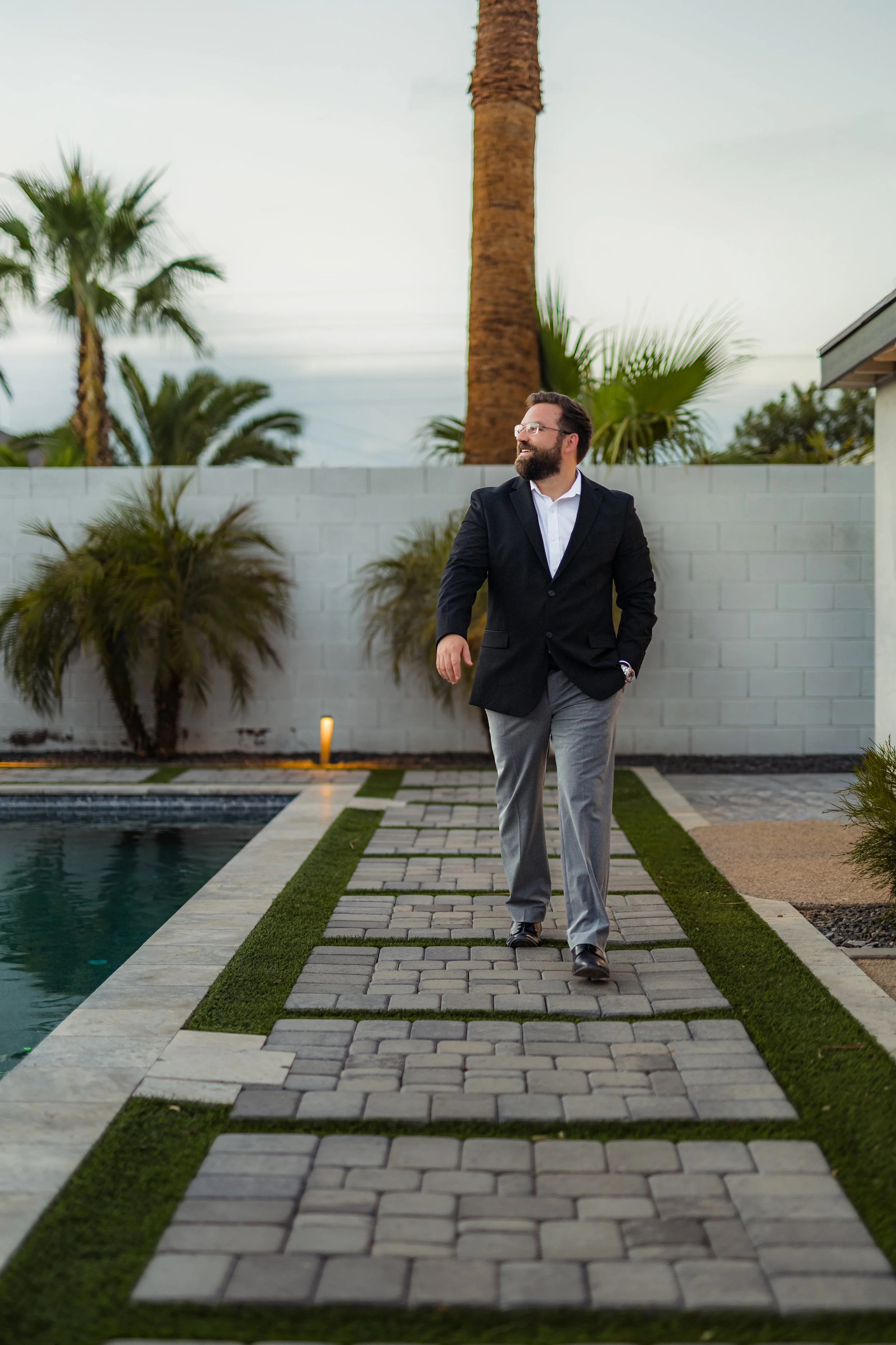 A man in a black blazer, white shirt, and grey trousers walking along a paved pathway next to a swimming pool, with palm trees and a white wall in the background, under an overcast sky.
