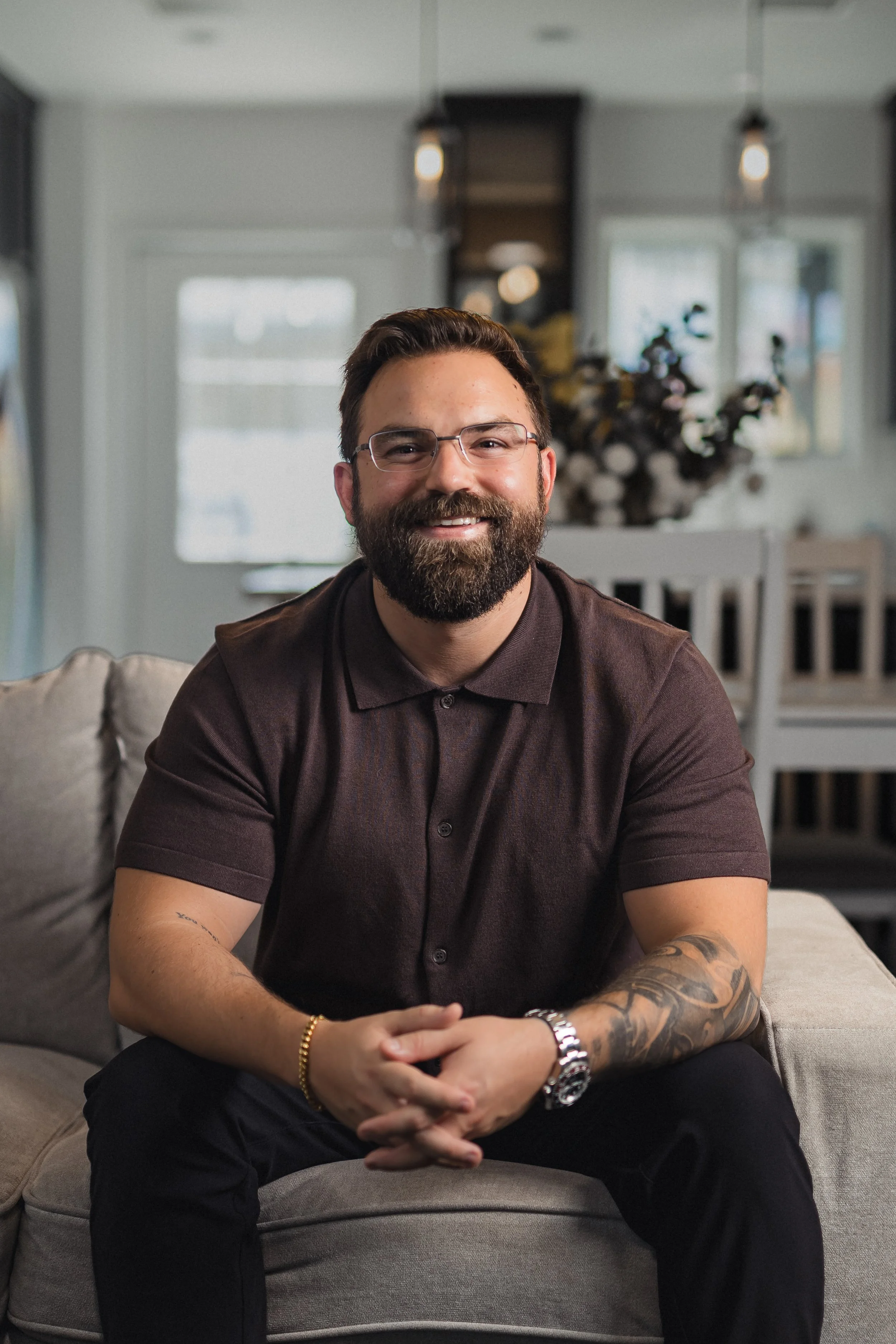 A smiling man with glasses, a beard, and tattoos on his right arm, sitting on a gray sofa in a living room with large windows and modern decor.