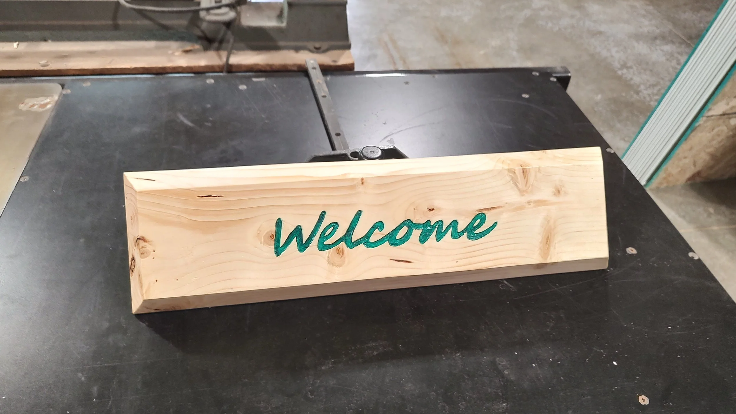A wooden welcome sign with green painted text 'Welcome' placed on a black workbench in a woodworking or workshop setting.