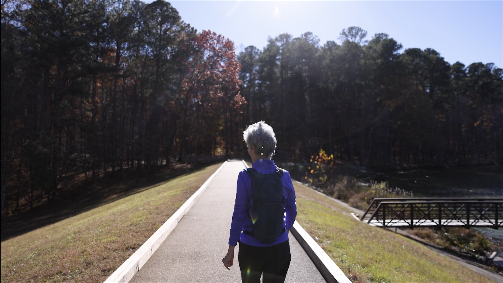 A woman with gray hair wearing a purple jacket and carrying a black backpack is walking on a paved trail in a forested park on a sunny day.