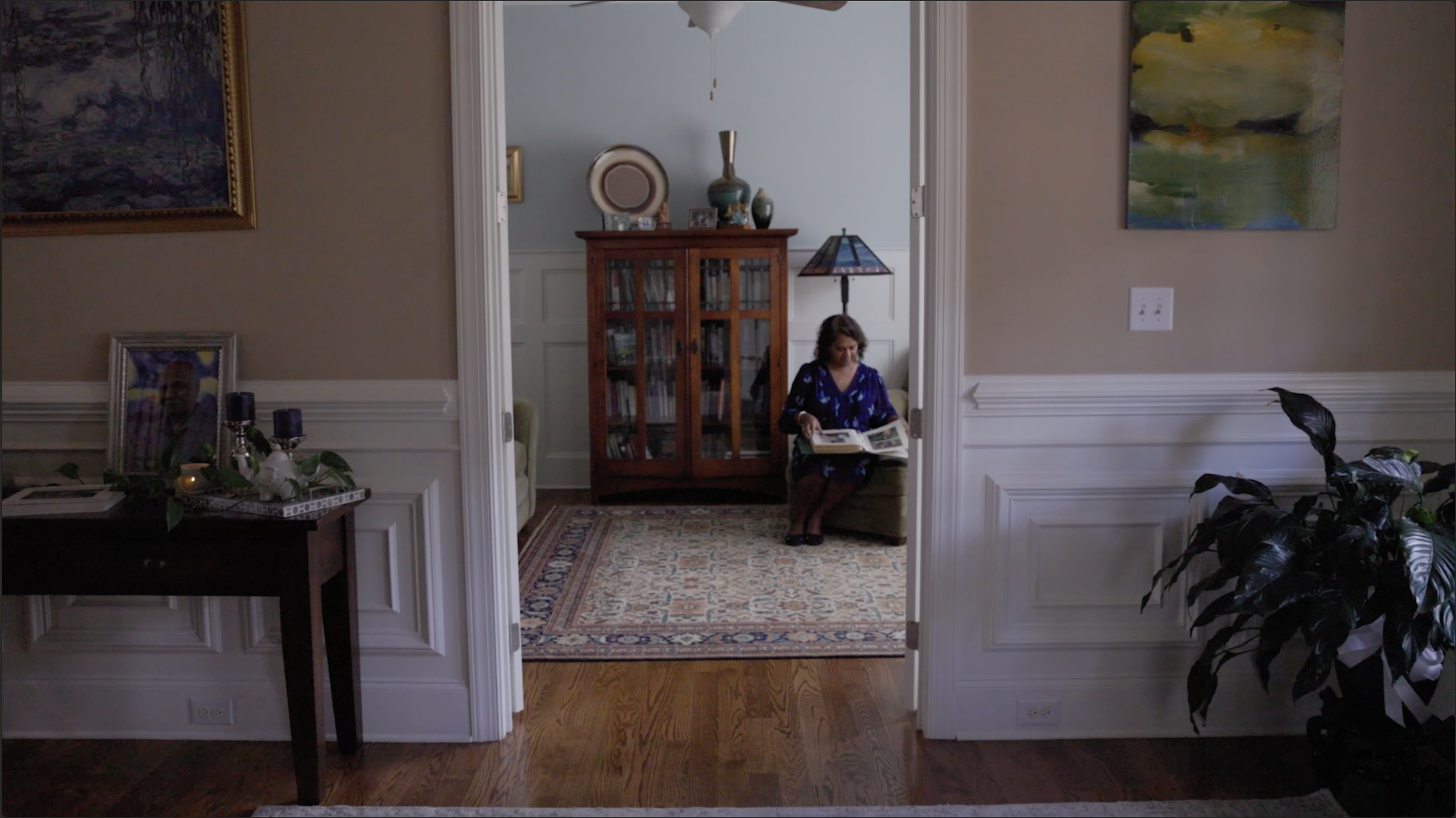 A woman in a blue dress sitting on a small armchair and reading a book in a living room, viewed through an open doorway. The room has artwork on the walls, a rug on the floor, and various decorative items such as vases and a lamp.