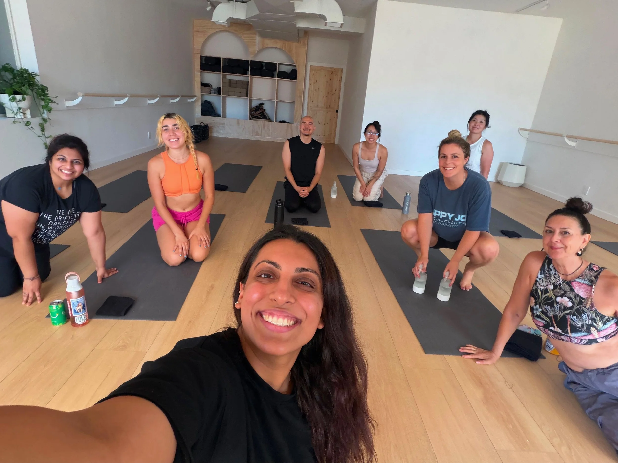A group of six women in a yoga studio sitting on yoga mats, smiling at the camera after a Joyflow class or gathering, with water bottles and drinks in front of them.