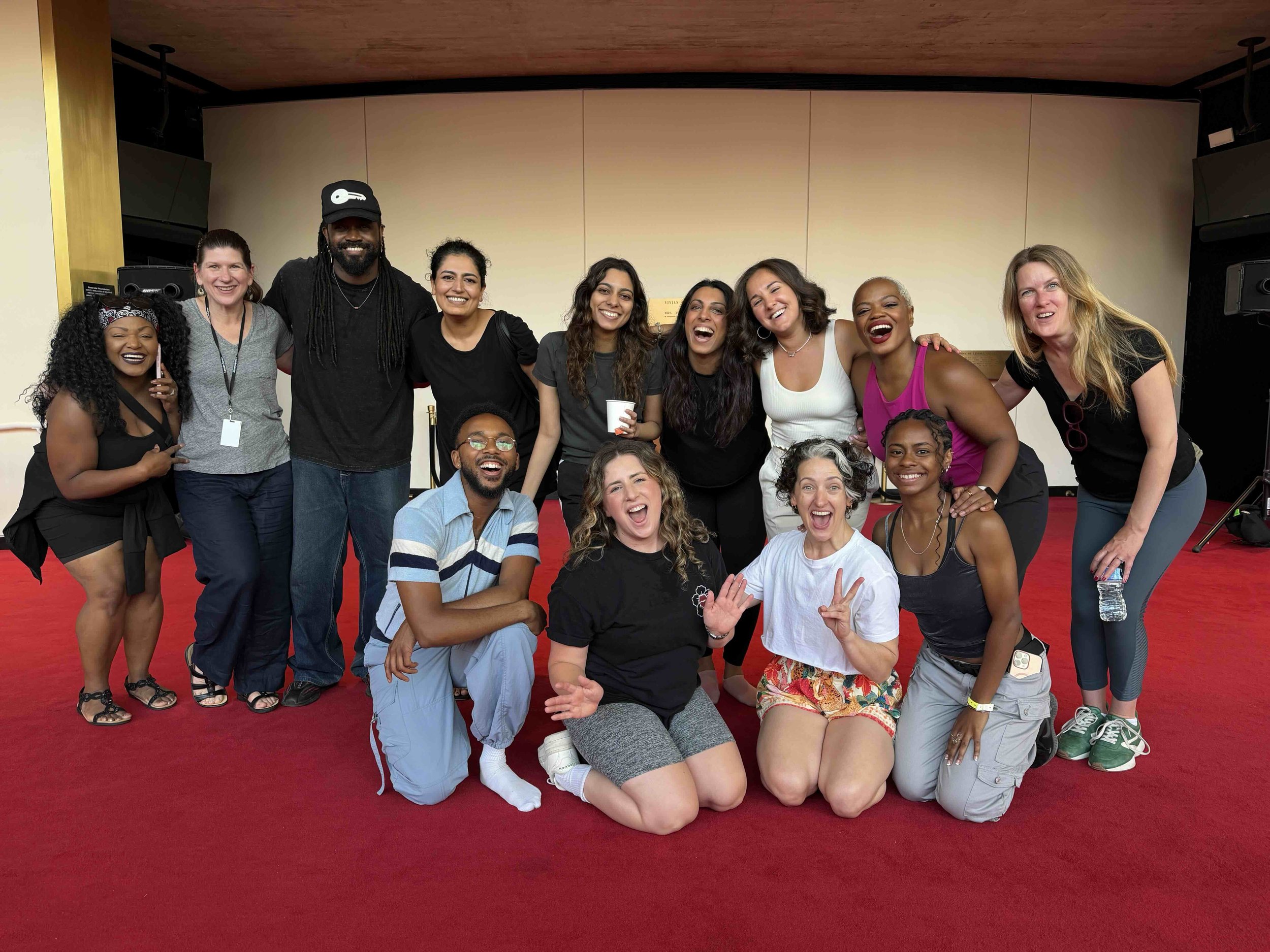 A group of twelve diverse people smiling and posing together on a red carpeted floor indoors, with a plain wall in the background.