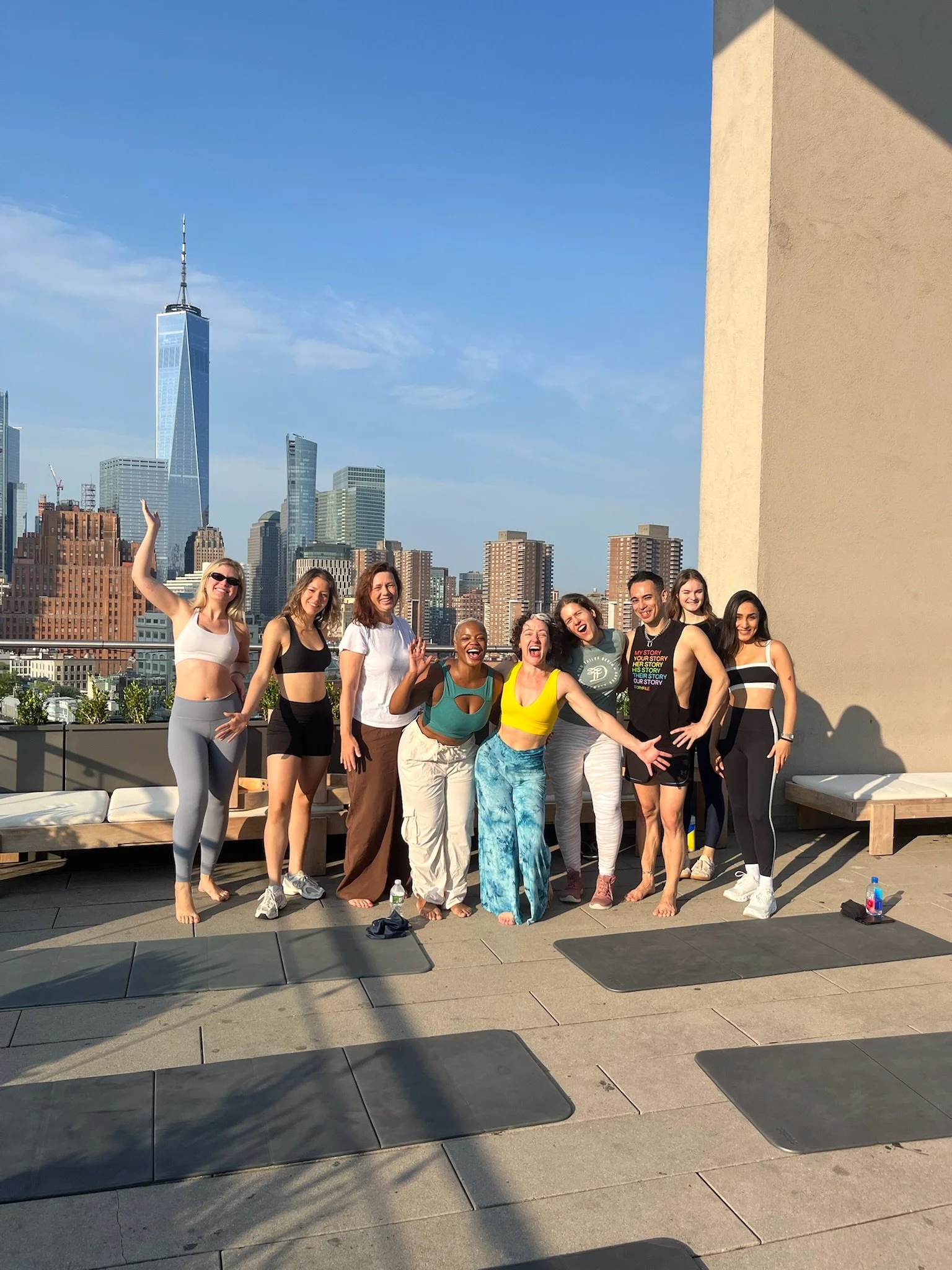 A group of ten diverse women standing on a rooftop with a city skyline, including One World Trade Center, in the background on a sunny day.