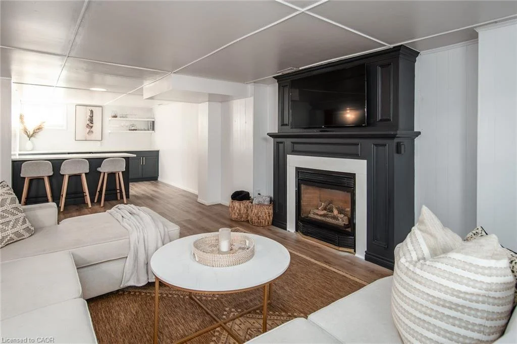 Contemporary living room with white sectional sofa, striped pillows, round white coffee table, fireplace with black mantel, and a flat-screen TV above it. In the background, a bar area with three gray stools, dark cabinetry, and a small shelf with de