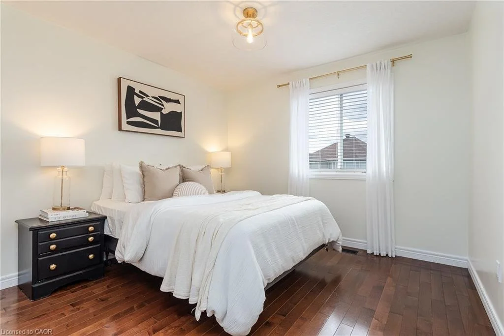 A cozy bedroom with a white bed and multiple pillows, black nightstands with lamps, a window with white curtains, hardwood floors, and a framed abstract art piece on the wall.