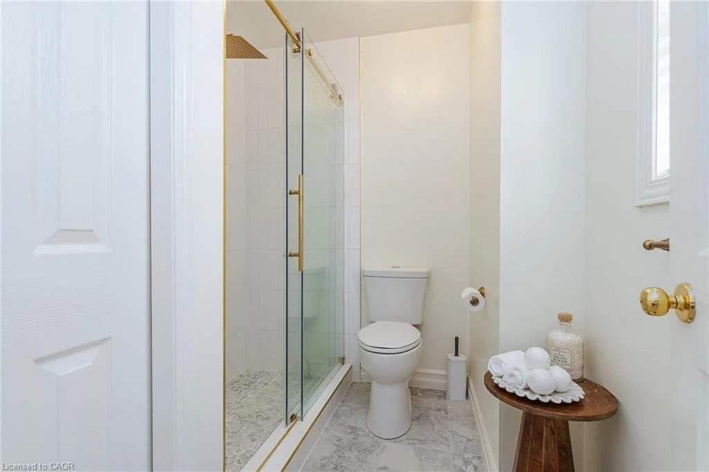 Small bathroom with a corner shower with glass doors, a white toilet, a small wooden side table with rolled white towels and a bottle, and a window with natural light.