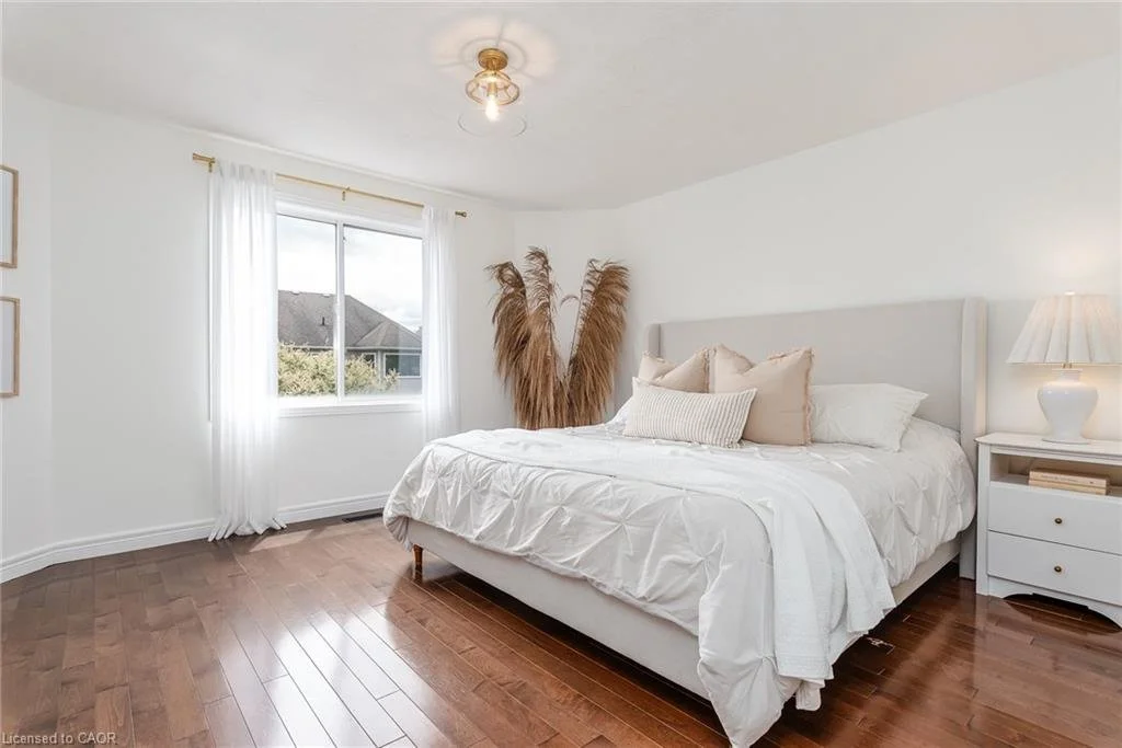 A bright bedroom with a large window, white curtains, a white bed with pillows, a nightstand with a lamp, and decorative pampas grass in the corner.