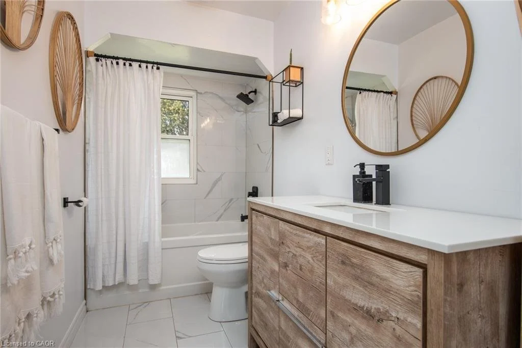 Modern bathroom with a wooden vanity, black fixtures, a large circular mirror, a shower with a marble wall, a small window, and white towels.