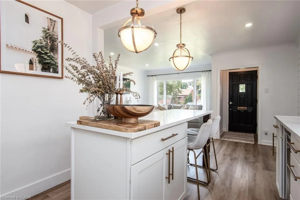 Kitchen with white cabinets, light wood flooring, black front door, and two hanging pendant lights. Includes a kitchen island with bar stools, a wooden bowl with dried branches, and a framed picture on the wall.