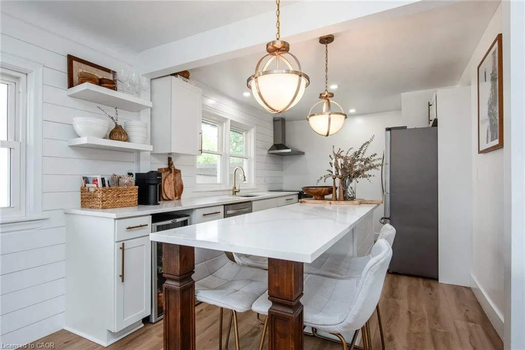 Kitchen with white cabinets and walls, a white countertop island with wooden support, two white cushioned chairs, pendant lights, a stainless steel refrigerator, open shelves with white bowls and decor, and a window with greenery outside.