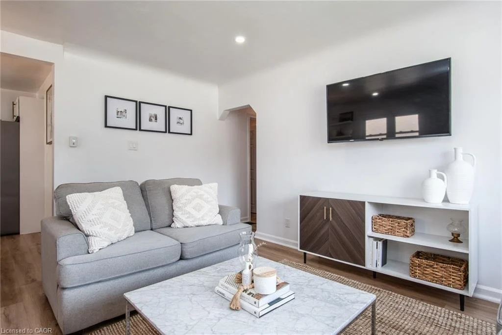 Living room with a gray sofa, white patterned pillows, a marble coffee table, wall-mounted flat-screen TV, and a white shelving unit with decorative vases and woven baskets.